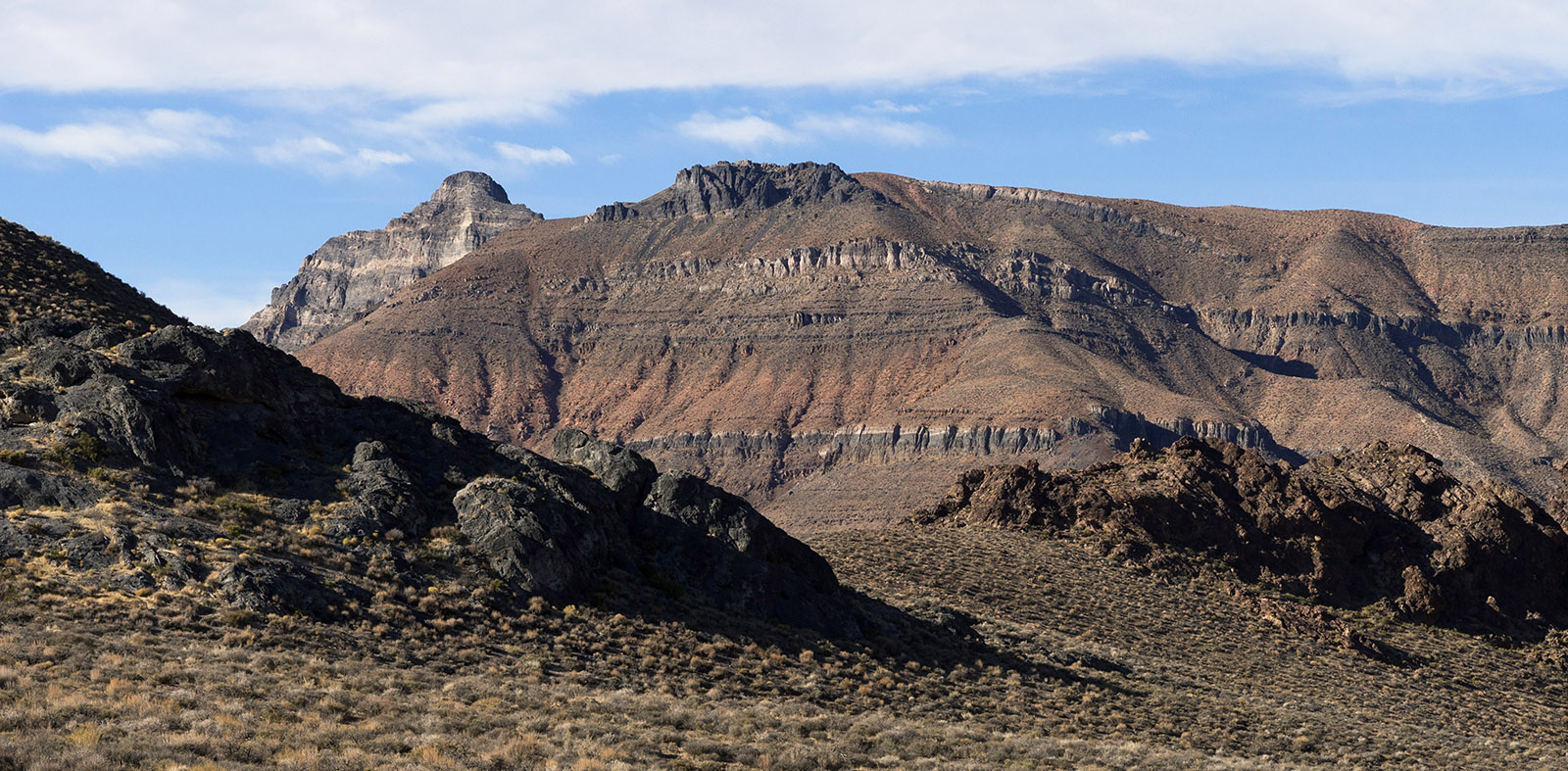 The highest peak on the left and to the rear is Thimble peak which is composed of Bonanza King Formation (Thick dolomite with thinner layers of limestone and shale from the mid to upper Cambrian Period, Paleozoic Era about 450 to 500 MA).  The wide mountain in front of Thimble Peak with horizontal strata is Carrera Formation shale and limestone from the mid to lower Cambrian period.  Foreground contains Tertiary volcanic rock on the left and Zabriskie Quartzite (lower Cambrian Period) on the right.