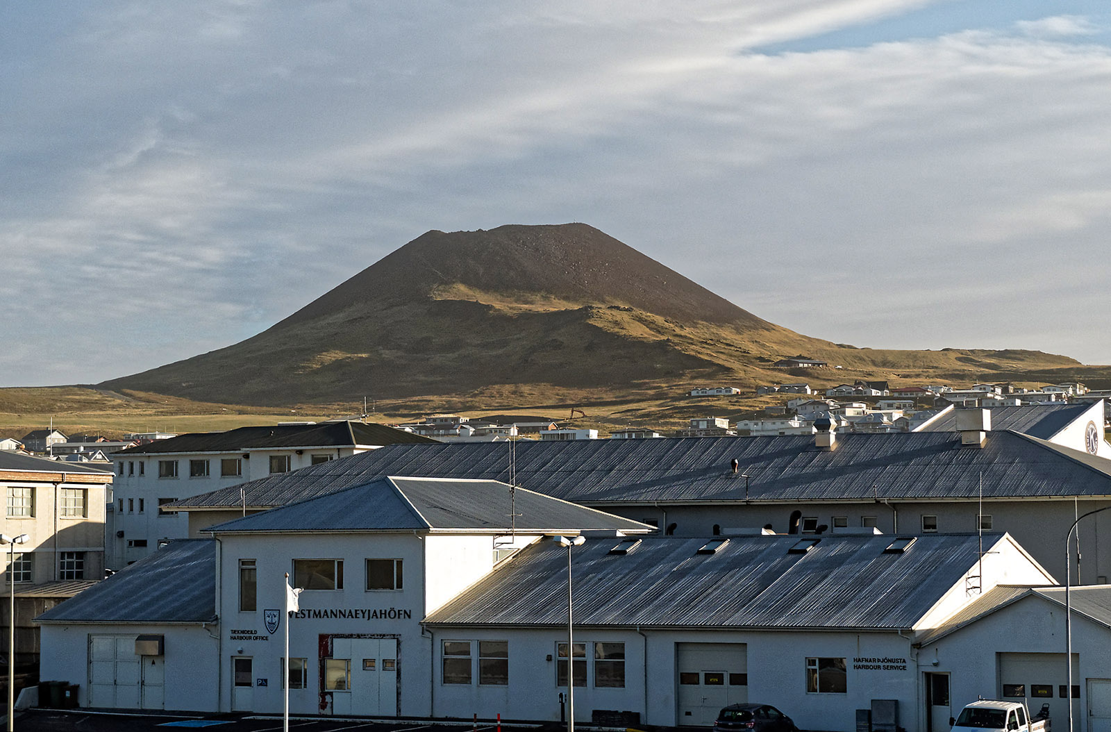 Eldfell is visible above the harbor buildings at Vestmannaeyjabaer. Eldfell is a volcano that was newly formed by an eruption in 1973 that forced the evacuation of the island and destroyed over 400 homes.