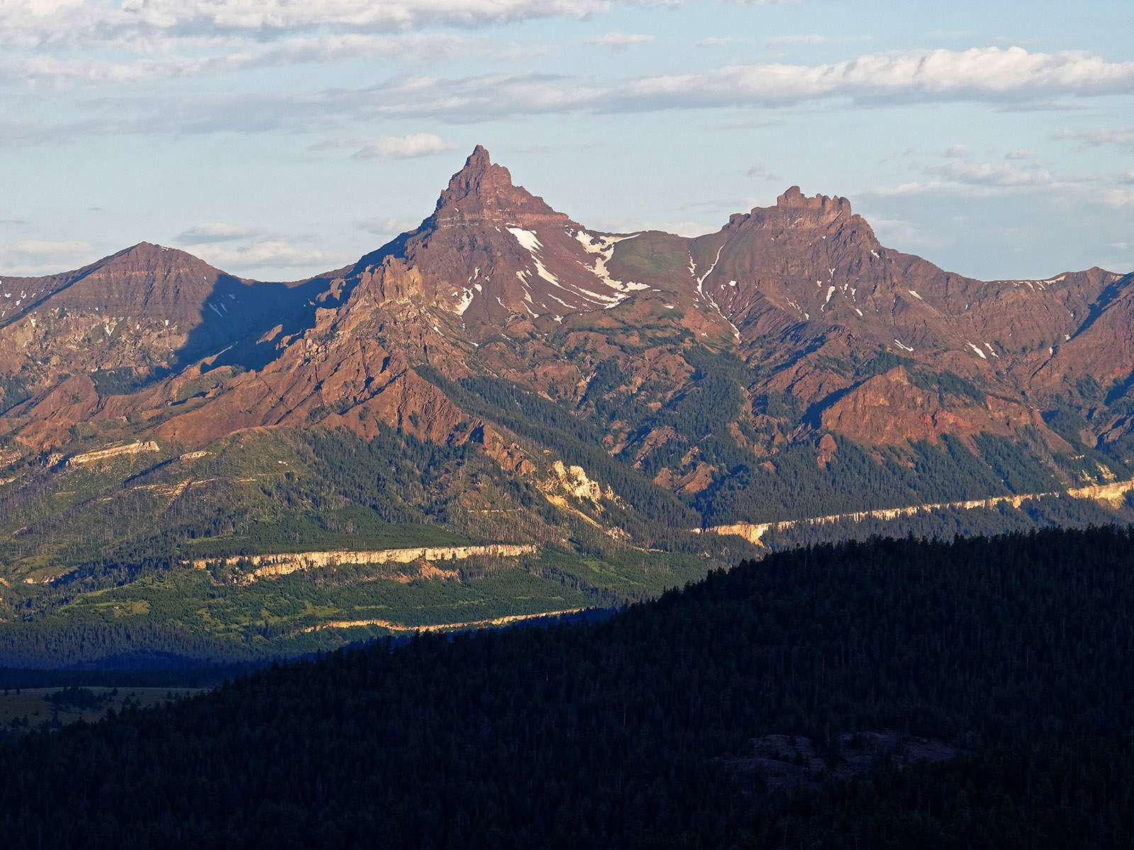 Pilot Peak (center) and Index Peak (right) are glacier-carved peaks of volcanic strata from Absaroka Volcanic activity that occurred 53 to 43.7 MA.  The Absaroka stratovolcanoes eroded and these peaks are eroded remains of Trout peak and Wapiti lava flows of andesite (lava of intermediate silica content).  The off white to light green line below the base of the peaks is the Pilgrim formation of limestone and shale from the late Cambrian period (550 Ma).